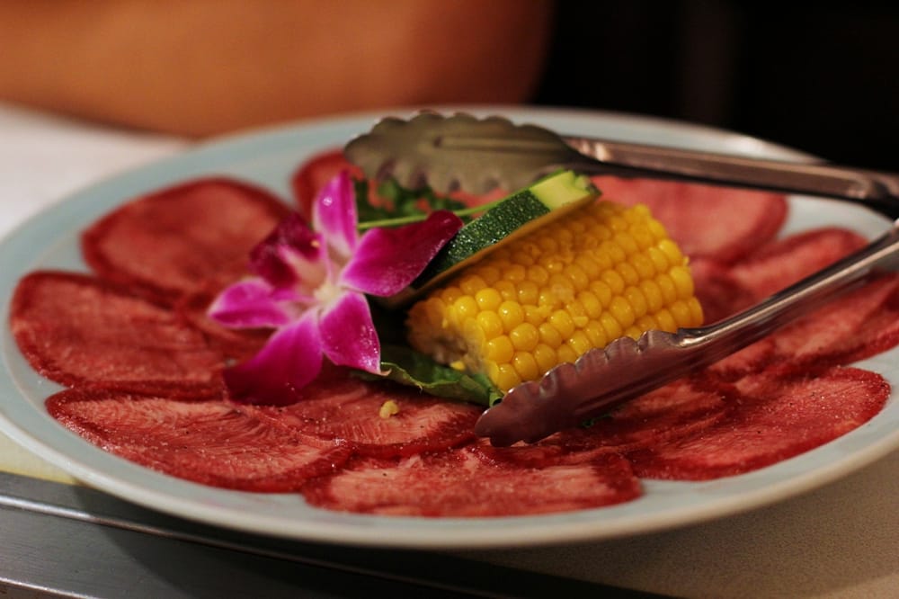 Thinly sliced salted beef tongue arranged on a plate with orchid garnish, corn, and tongs